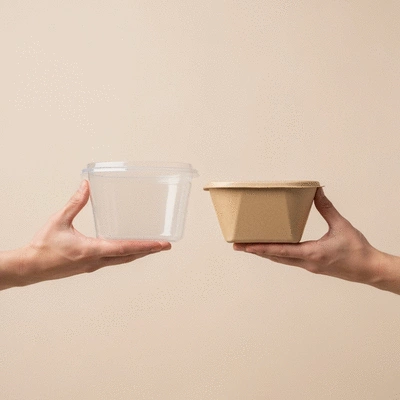 Two hands holding a compostable food container and a plastic food container, side by side, on a neutral background
